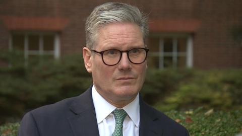 The Prime Minister Sir Keir Starmer in a dark suit an white shirt with a green tie, wearing black-framed glasses, standing in the Downing Street garden 