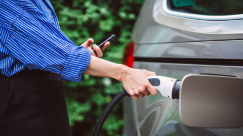 A hand holding an EV charger at a silver car. The person is wearing a striped blue and white shirt, and black trousers