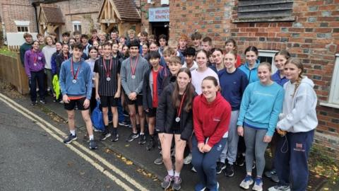 A large group of teenagers pose outside the front entrance of their rowing club, squeezed onto a narrow pavement. Many of them are wearing medals and smiling for the camera. 