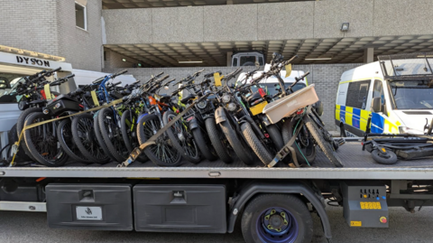 Recovery truck with e-bikes on the back of it. It is a large vehicle and parked in front of a Devon and Cornwall police van.