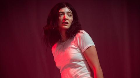 Lorde on stage at Glastonbury. The singer, lit in a red light, wears a white T-shirt, her arms by her side. Her dark hair is worn loose and messy. She looks over her left shoulder towards the crowd. 