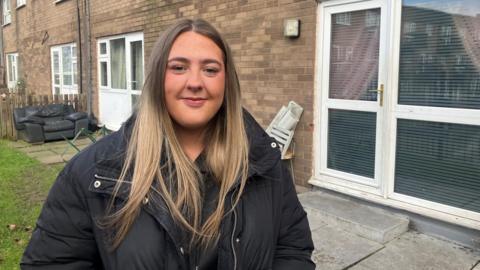 Kelsey Schofield, 18, who has long straight blonde hair and is wearing a thick black coat, smiles at the camera while standing on a lawn outside a ground floor flat. 