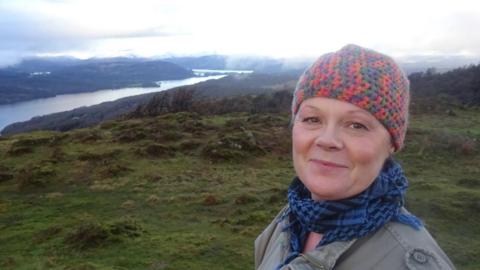 Cat, a woman wearing a red and grey woolly hat and blue scarf, she smiles as she stands in a field, you can see a large lake in the background.
