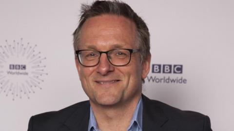 Michael Mosley, wearing a dark suit and blue shirt, smiles in a press photograph. He has short dark grey hair and is wearing black-rimmed glasses.