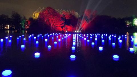 A light display with lots of small blue lights floating on a lake. The stadium St James' Park is peaking out from behind the trees which are lit up by a red light.
