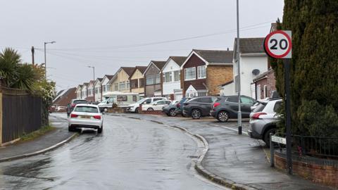 The entrance to a very wet residential street on a rainy, grey day. Houses can be seen on the right-hand side of the road, which have driveways with parked cars on. On the left-hand side, small palm trees can be seen behind a garden fence and there is a white Audi parked on the road, slightly mounting the pavement.