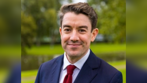 Alan Strickland, a white man with brown hair, wearing a navy blue suit and red tie, smiling