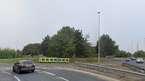 A roundabout with a neon arrow sign in its centre. There is one car approaching the roundabout and three cars coming off the roundabout and driving on the opposite carriageway. 