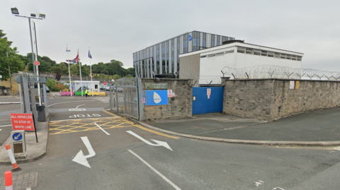 A image of the front entrance to the current Babcock building in Plymouth. It is a large glass building with a brick entrance and security gates. There are cars parked outside the front of the building.