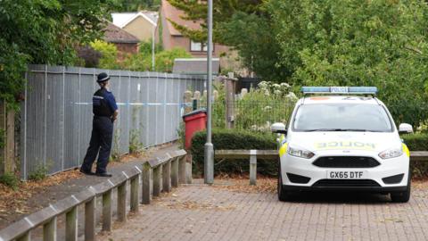 An alleyway taped off with blue and white police tape. A police officer stands at the edge of the tape cordon. A police car is parked next to the cordon.