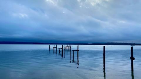 An idyllic image of the sea stretching out far ahead. The sky is filled with dark, threatening clouds. Hills can be seen in the far distance. There are wooden struts coming out of the sea in a line.