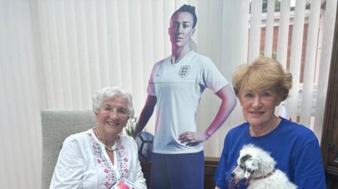 Gloria Stapylton (left) has short white hair and is smiling. She wears a pearl necklace and white cardigan. She is holding a copy of Women's Health magazine, which has a picture of Lucy Bronze on it. Julie Tough (right) has short ginger hair and holds a small white dog and a newspaper. A cardboard cutout of Lucy is behind them.
