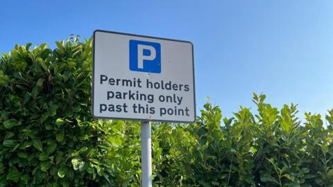 A white sign has a blue P on it with 'permit holders parking only past this point' written on it. It has been put up in front of a thick hedge.