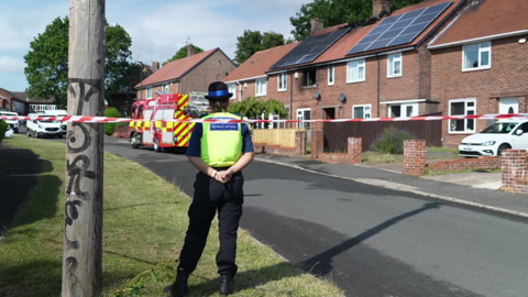 A police officer stands with their back to the camera next to police tape. The damaged terraced house is in the background. There is smoke damage to an upstairs window.
