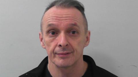 A pink-faced man wearing a black top and with a receding hairline stares at the camera.