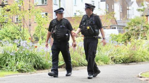 Two police officers walk around a park in Exmouth during a sunny day with lots of plants behind them in flowerbeds.