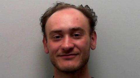 A custody picture of a young man with short, thinning curly hair, who sits against a plain light grey wall, wearing a grey crewneck sweatshirt. He looks directly at the camera with a relaxed posture and a subtle smile.
