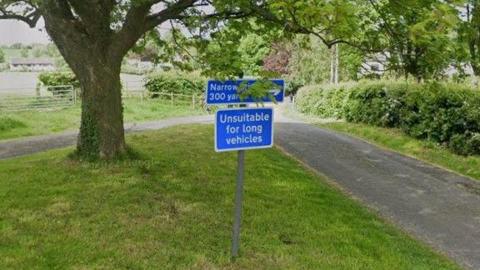 A country  lane runs along the right of the picture while on the left a blue road sign reading "unsuitable for long vehicles" can be seen on a grass verge under a tree.