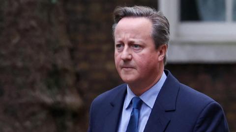 Lord David Cameron wearing a blue suit, blue shirt and blue tie. He is walking and has a serious expression