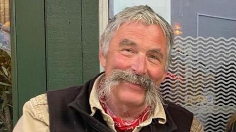 A head and shoulder photograph of David Oliver from Marlborough smiling with grey hair and a large grey moustache wearing a pale yellow shirt, a brown waistcoat and a red and white neckerchief, with a green glass door in the background. 