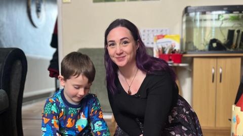 Jace wearing a blue sweatshirt with colourful graphics and black trousers sitting next to Sarah wearing a black shirt and a black and pink floral skirt playing with logos