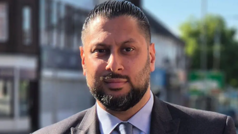 A man facing the camera. He has black hair and a black beard and moustache. He is wearing a grey suit with a blue shirt and tie.