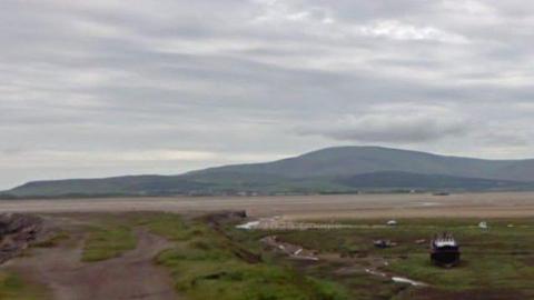 A Google Streetview showing the tide out at Duddon Estuary. There is a boat on a grassy area of dry land. Beyond that is a huge stretch of sand with a line of water - akin to a meandering stream - but the tide is a long way out of shot to the left. Beyond the wide estuary, in the background, hills gradually rise up from the left to a domed peak, creating what looks from distance like a rolling ridge. There are plenty of grey clouds but they are high and the day appears dry.
