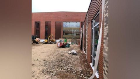 A red brick building with large windows has dirt on the ground in front of it, with a yellow digger, a skip and construction equipment.