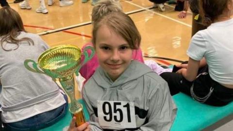 A young girl, her hair tied back, sits on a bench with other girls similar in age to herself. She is holding a trophy.