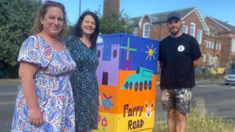 Two women and a man standing next to a utility box painted in bright colours with a windmill, train and "poppy road" written on it.