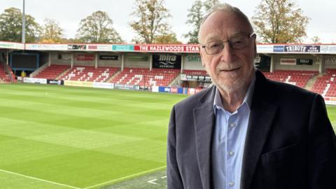 Adrian, who wears square-framed glasses, a blue shirt and a navy suit jacket smiles at the camera from inside the empty Cheltenham Town FC stadium.