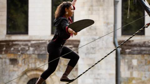 A woman in black leggings and a black hooded top climbing up a high wire while holding a fan.