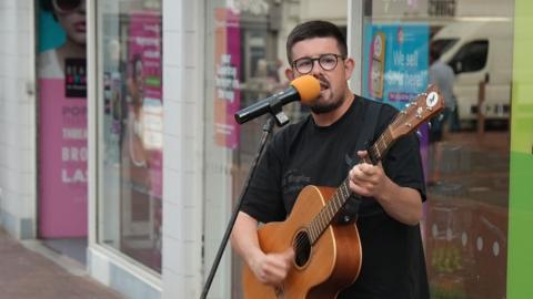 A male musician with dark hair, a cropped beard, glasses and a black T-shirt, sings into a microphone while playing his acoustic guitar in front of a shop window.