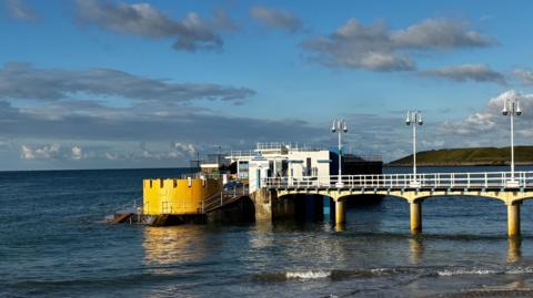 The Havre des Pas lido at high tide, it is sunny with some clouds in the sky. 