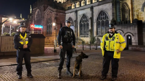 Three male police officers stand in a street at night. One officer's black uniform is covered with a florescent vest. A second is wearing all black and has a dog on a lead next to him. The third officer has a florescent jacket and vest one with dark trousers.