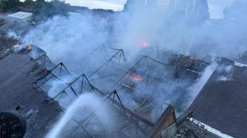 A large warehouse roof on fire as it is hosed down from above
