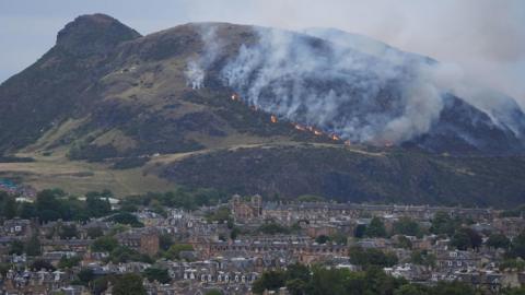 A view of a wildfire on Arthur's Seat in Edinburgh. Large amounts of smoke can be seen coming off the middle of the hill, where there are also specks of orange flames. The sky is grey. The city is in the foreground.