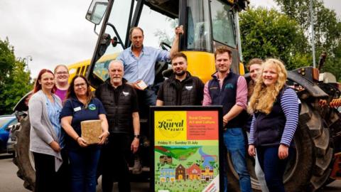 Five women and four men stood around a yellow tractor alongside a colourful sign which reads The Rural Remix