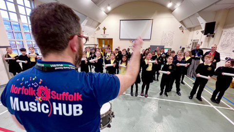 A man in a blue T-shirt with Norfolk & Suffolk Music Hub across the back holds out a drumstick towards a school hall of primary school children holding musical shakers