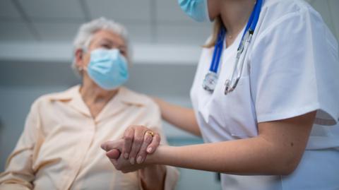 An elderly woman with grey hair wearing a face mask has her hand held by a female doctor also wearing a mask