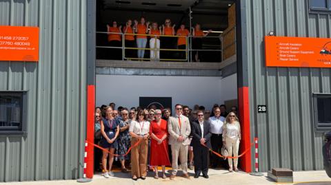 A group of people from the Needles and Pins team standing behind red ribbon at the opening of an industrial steel building.