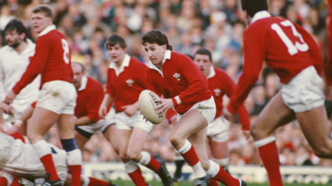 Jonathan "Jiffy" Davies is holding a rugby ball while playing in an international match for Wales. He is wearing a red Wales jersey, white shorts and red socks. Surrounding him are players also wearing the same kit, and a crowd of spectators are sat down, blurred, in the distance.