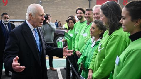 King Charles III speaks to members of staff who are all in green jumpers and are smiling as he speaks