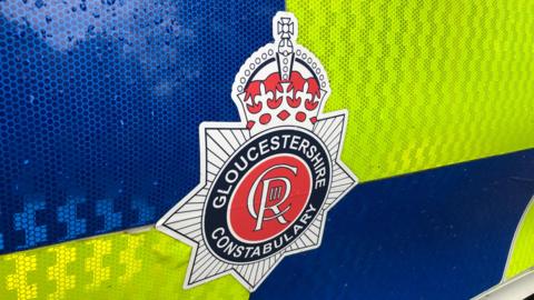 A close up of the Gloucestershire Constabulary badge on a police car door which has blue and yellow hi-vis chequering. The badge includes the cypher of King Charles III emblem (a stylised CRIII) and a crown.