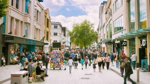 Cardiff's Queen Street which is pedestrianised with lots of people walking along, a big tree in the middle and shops on either side