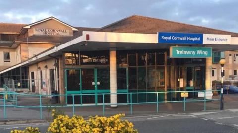 The entrance to the Royal Cornwall Hospital. There is a sign for "main entrance" and the "Trelawny Wing". There are green doorframes and railings in front of the building.