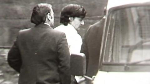 A black and white still from TV footage showing Peter Sullivan, who has thick black hair and is wearing a white shirt, being led to a police van in handcuffs.