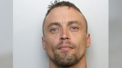 Custody picture of Ashley Billing. He is staring directly into the camera, and has short light brown hair and stubble. He is standing against a plain grey background and a neck tattoo is faintly visible on the right side of his neck. 