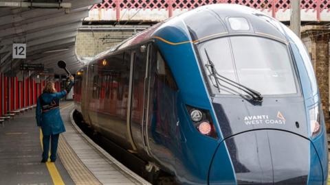 An Avanti West Coast staff member stands next to a blue Avanti West Coast train at a station. 