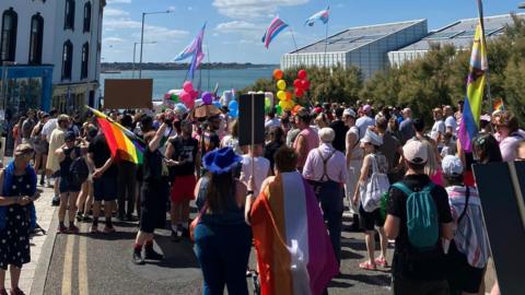 A crowd of colourfully dresses people waving flags walking away from camera towards the seafront for Margate Pride 2025